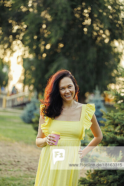 Glückliche Frau im gelben Kleid mit Kaffeetasse steht im Park