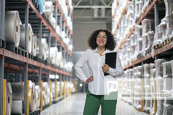 Happy businesswoman with tablet PC standing by rack at warehouse