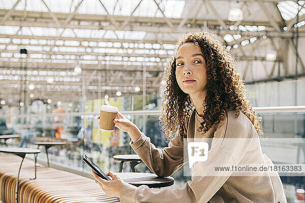 Young woman sitting with mobile phone drinking coffee at train station