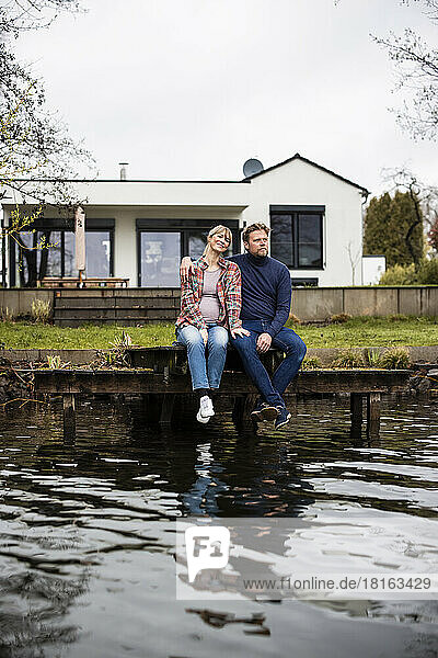 Expectant couple sitting together on jetty at lake in front of house