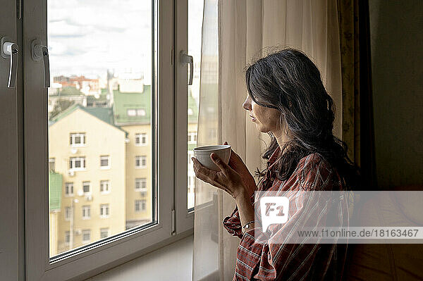 Frau hält Kaffeetasse in der Hand und schaut zu Hause durch das Fenster