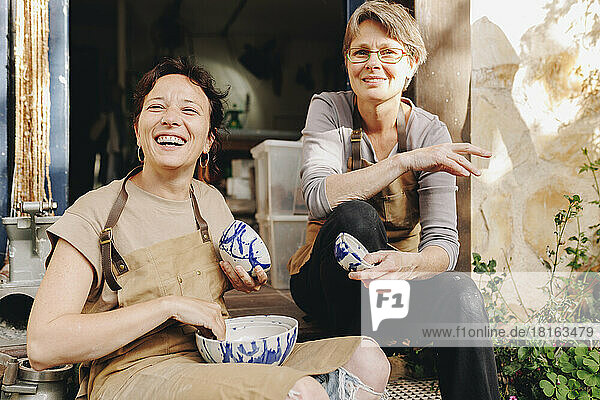 Cheerful entrepreneur with colleague holding ceramics bowls sitting in doorway