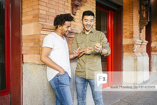Young multiracial men laughing and using phone together on footpath