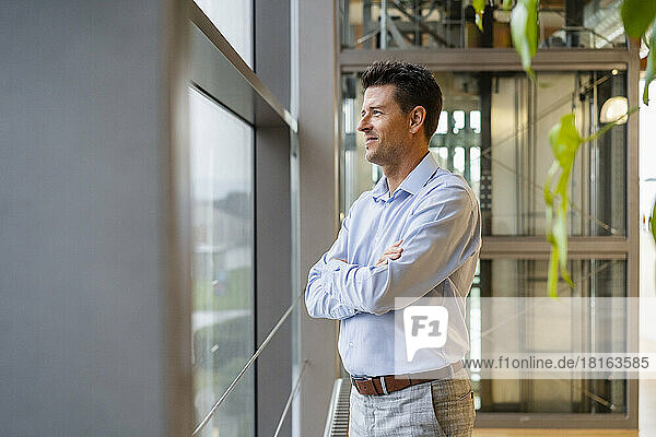 Smiling thoughtful businessman with arms crossed looking through window in office