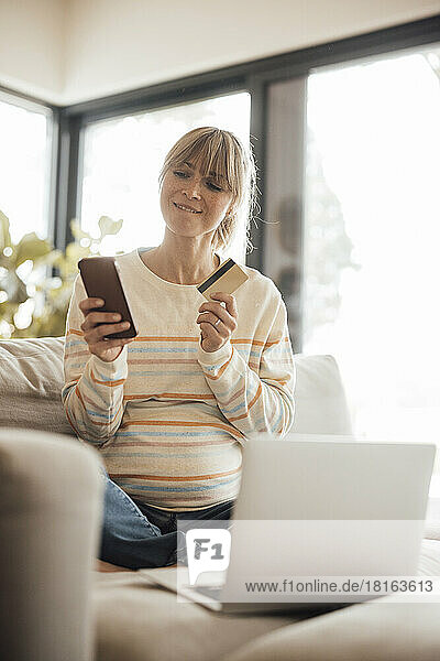 Pregnant woman holding credit card and smart phone doing online shopping at home