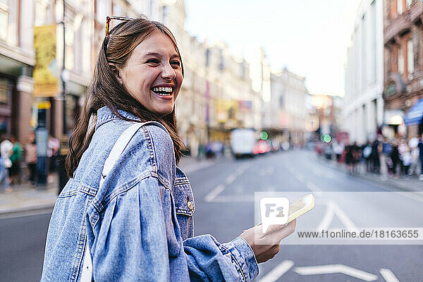 Fröhliche junge Frau mit Mobiltelefon spaziert durch die Stadt
