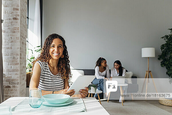 Smiling woman holding tablet PC with friends in background at home