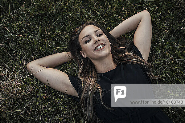 Happy young woman with hands behind head relaxing on grass at park