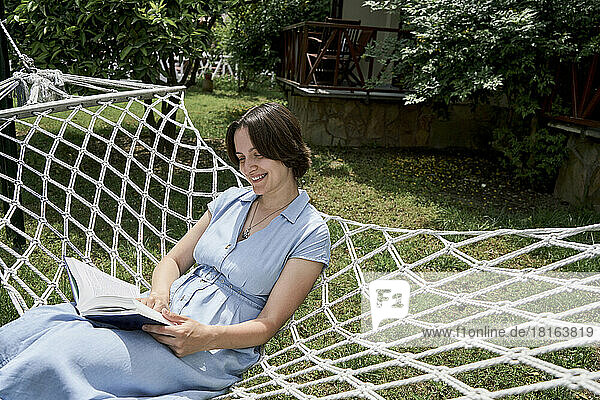 Smiling woman reading book sitting on hammock at sunny day
