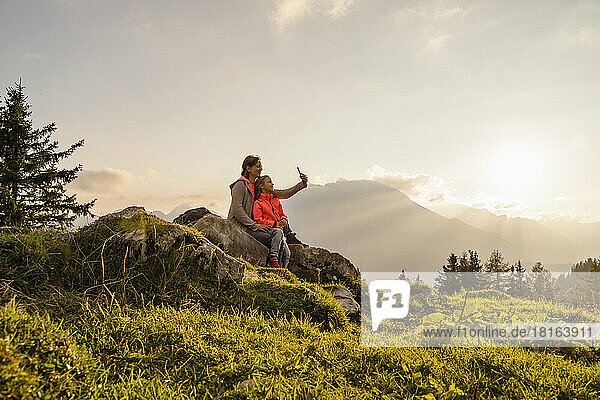 Smiling mother with daughter taking selfie through mobile phone sitting on rock