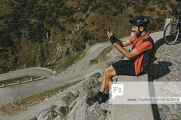 Cyclist photographing through smart phone sitting on wall at Gotthard Pass