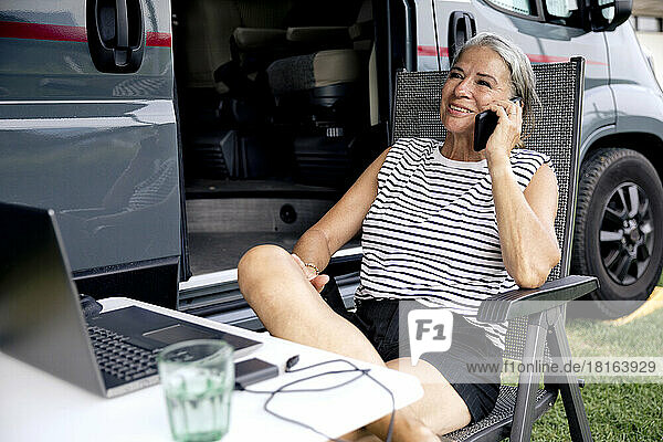 Smiling freelancer talking on mobile phone sitting outside camper van