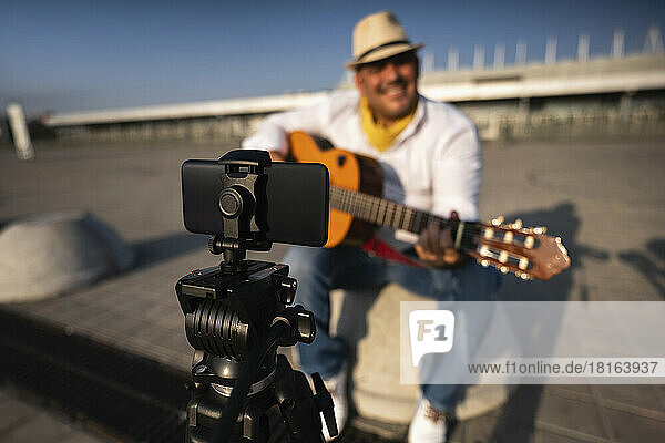 Street musician playing guitar and recording through mobile phone on tripod