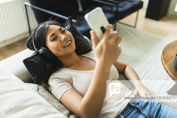 Happy woman wearing wireless headphones and using smart phone on sofa at home