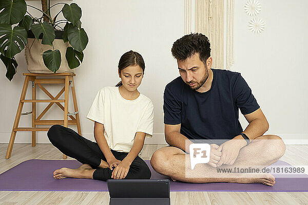 Man with daughter watching yoga tutorial on tablet PC sitting on exercise mat