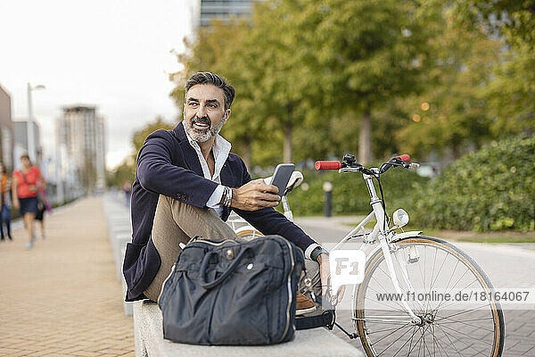 Happy businessman with smart phone resting on bench by bicycle