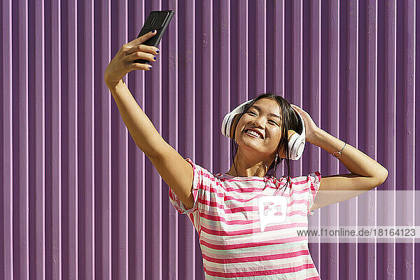 Lächelnde junge Frau macht ein Selfie mit ihrem Smartphone vor einer Wellblechwand