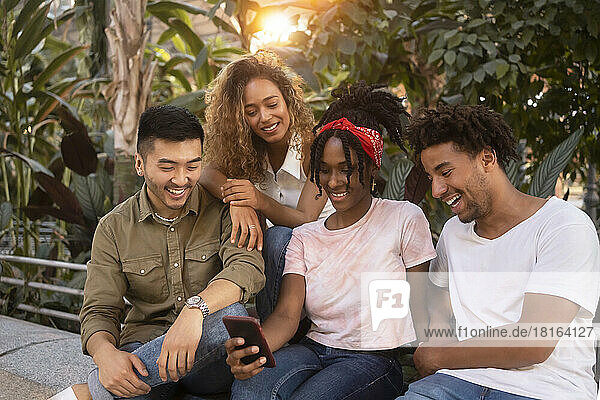 Young woman sharing smart phone with happy friends sitting on wall