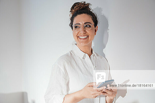 Smiling thoughtful woman with mobile phone by wall at home