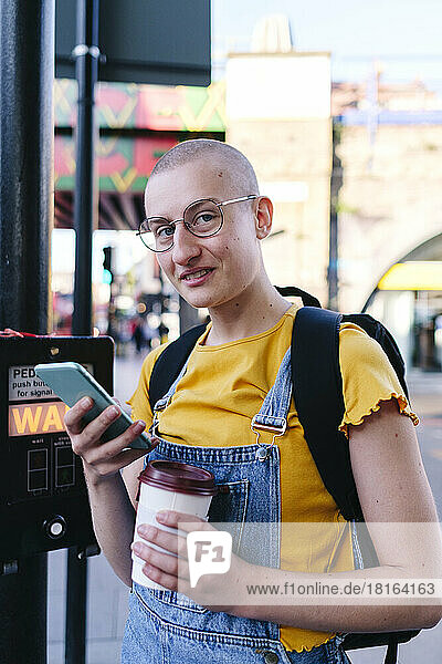 Non-binary person with eyeglasses holding disposable coffee and mobile phone