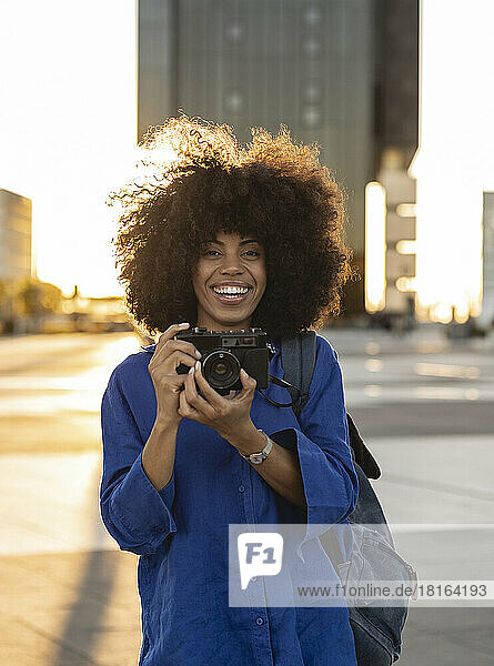 Cheerful Afro woman holding camera on footpath