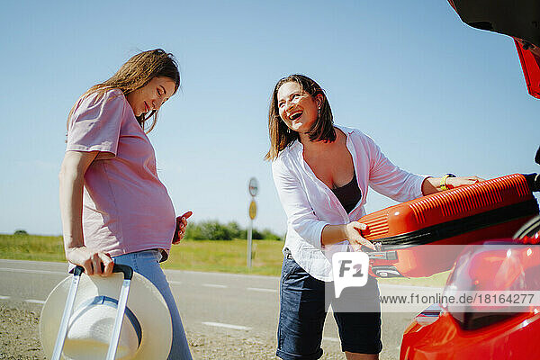 Happy woman loading suitcase in car trunk and looking at pregnant friend