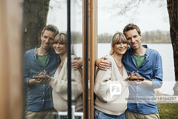 Smiling expectant parents holding baby booties by glass reflection