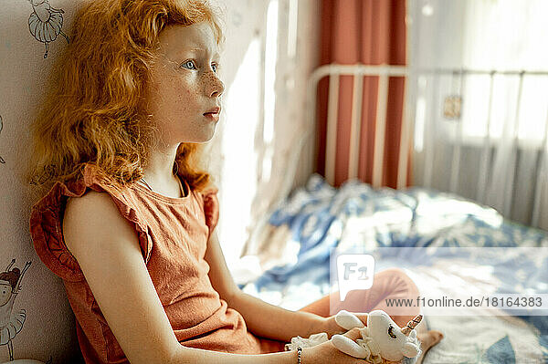 Thoughtful redhead girl with soft toy sitting on bed