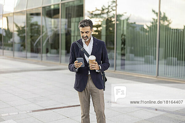Businessman with disposable cup and mobile phone standing in front of office building