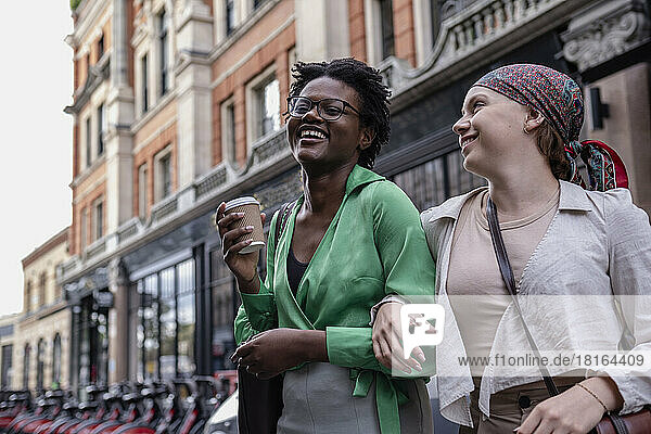 Happy woman holding disposable cup with friend wearing bandana in front of building