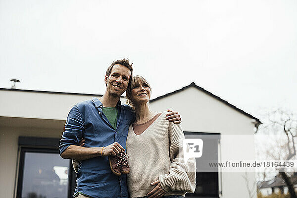 Happy expectant couple with arm around each other in front of house