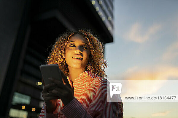 Smiling woman with mobile phone contemplating at sunset
