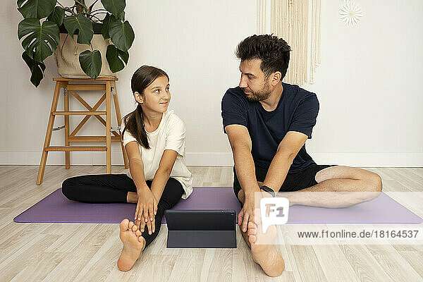 Man with daughter exercising yoga by looking at each other in front of tablet PC