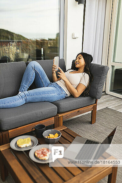 Glückliche junge Frau benutzt Smartphone auf dem Sofa im Wohnzimmer