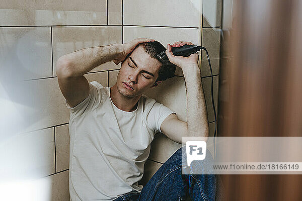 Depressed young man sitting with eyes closed shaving hair