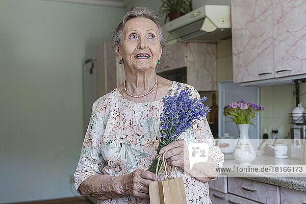 Happy senior woman with bunch of lavender flowers day dreaming in kitchen at home