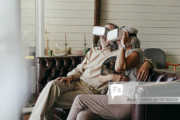 Happy couple wearing virtual reality simulator glasses embracing cat at home