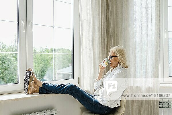 Relaxed senior woman drinking tea sitting with feet up near window at home