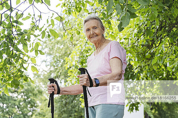 Lächelnde ältere Frau mit Wanderstock steht vor Baum