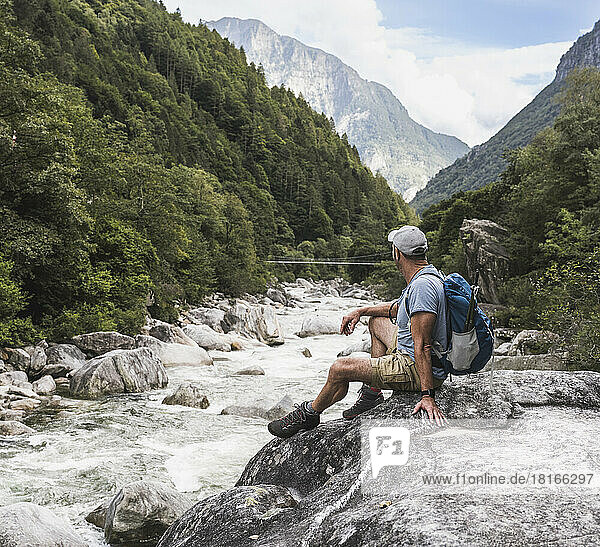 Reifer Mann mit Rucksack sitzt auf einem Felsen am Fluss