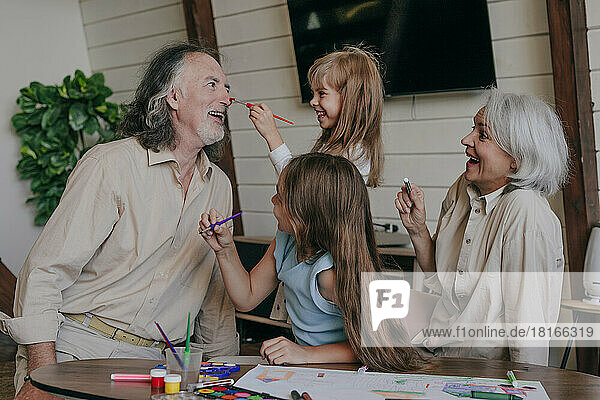 Granddaughter having fun painting grandfather's nose at home