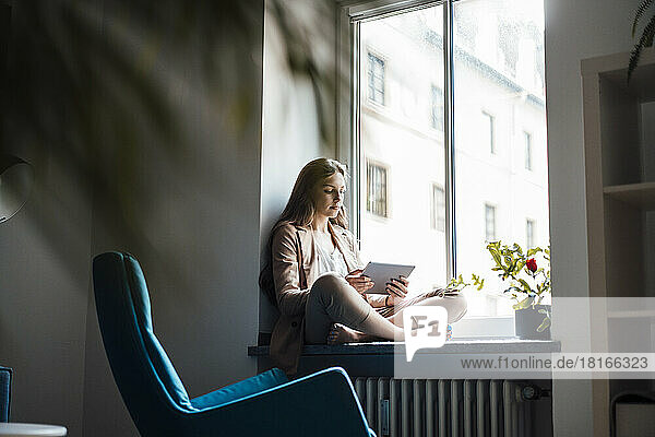 Businesswoman using tablet PC sitting on window sill at home
