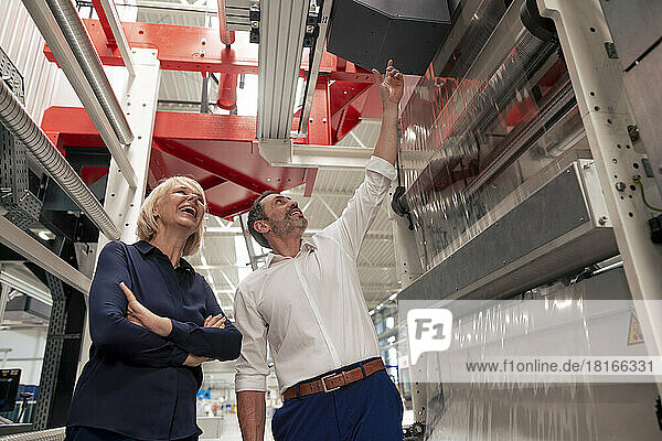 Smiling businessman pointing at machine to businesswoman with arms crossed in industry