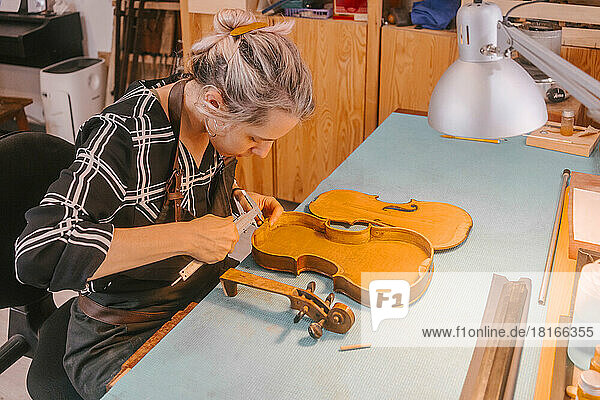 Instrument maker with tool carving and repairing violin at desk in workshop
