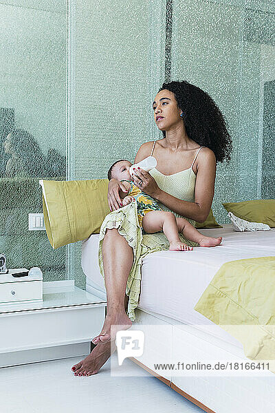 Woman feeding baby boy with bottle sitting on bed at home