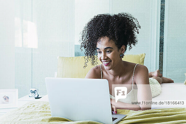 Happy woman with curly hair lying on bed using laptop at home