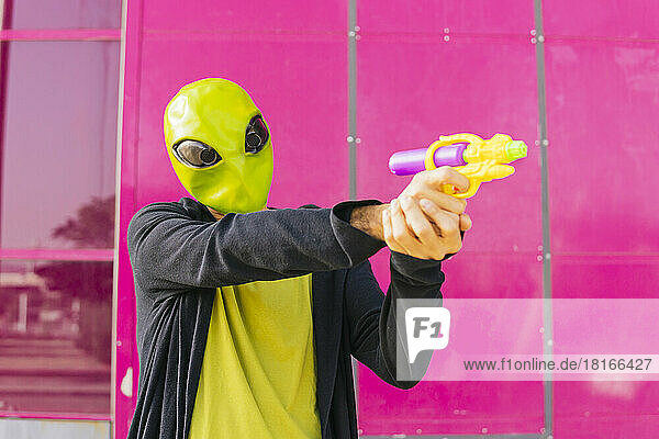 Mann mit Alien-Maske spielt mit Spielzeugpistole vor rosa Wand