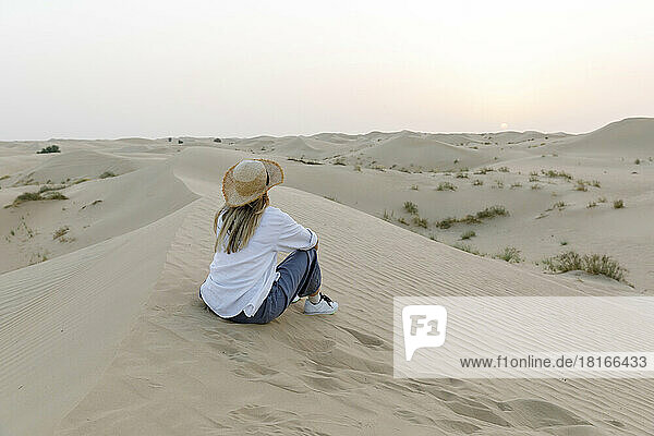Woman wearing hat sitting on sand in desert at sunset