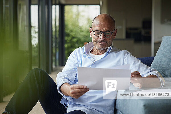 Senior man reading documents sitting by sofa at home