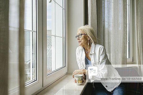 Ältere Frau mit Tasse schaut zu Hause aus dem Fenster